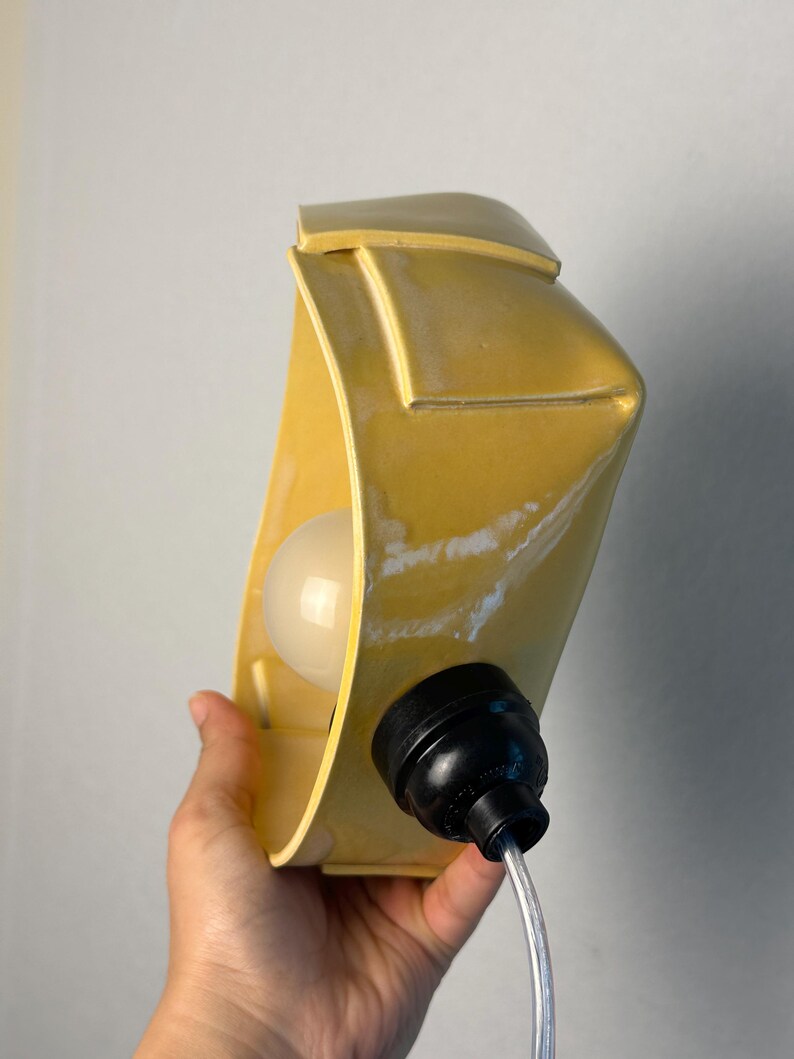 May include: A yellow ceramic wall lamp with a curved, rectangular form and a smooth, glossy finish. The fixture features a central white light bulb socket and a black plastic electrical component attached to the side with a clear power cord.