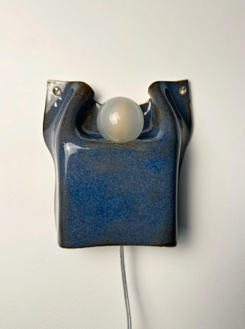 May include: A wall-mounted ceramic lamp featuring a deep blue, glossy glaze with a textured finish. The fixture has a curved, organic shape with two mounting holes at the top corners and a single, spherical, frosted white glass light bulb centered.