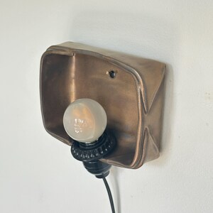 May include: A bronze-colored wall sconce with a rectangular, slightly curved shade. A round, frosted light bulb is set in a black socket. The sconce is mounted on a white wall, with a black electrical cord extending downward.