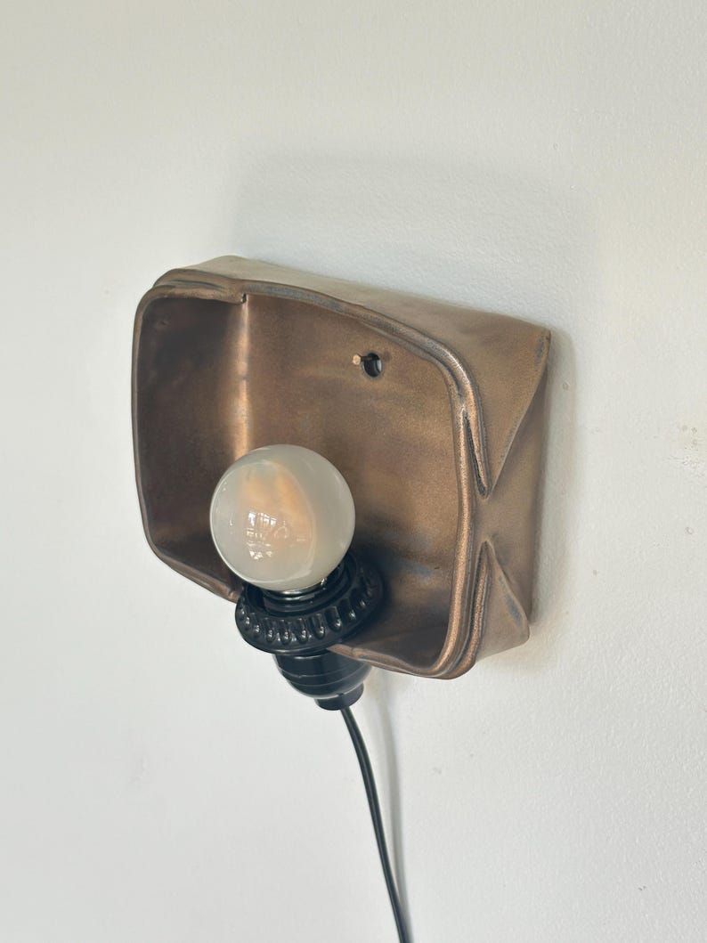 May include: A bronze-colored wall sconce with a rectangular, slightly curved shade. A round, frosted light bulb is set in a black socket. The sconce is mounted on a white wall, with a black electrical cord extending downward.