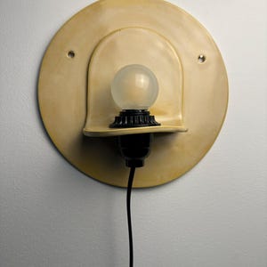 Pu&ograve; includere: Una lampada da parete con una piastra posteriore circolare in ceramica giallo pallido dotata di due piccoli fori. Un portalampada nero sostiene una lampadina rotonda in vetro smerigliato, posizionata su un piccolo ripiano curvo in ceramica.