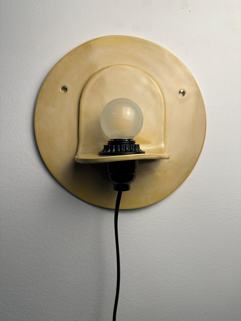 Pu&ograve; includere: Una lampada da parete con una piastra posteriore circolare in ceramica giallo pallido dotata di due piccoli fori. Un portalampada nero sostiene una lampadina rotonda in vetro smerigliato, posizionata su un piccolo ripiano curvo in ceramica.