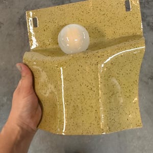 May include: A wall-mounted ceramic light fixture with a glossy, speckled mustard yellow glaze. The rectangular base features two rectangular cutouts at the top for mounting and a central circular opening holding a small, glowing white glass bulb.