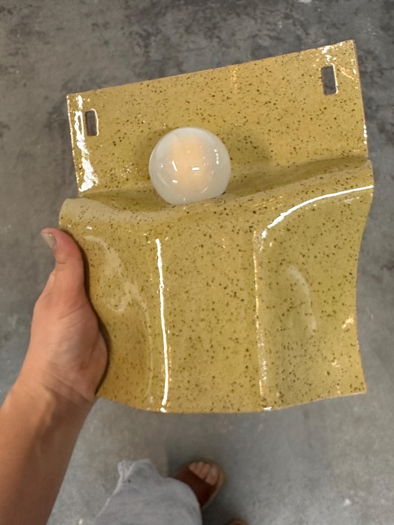 May include: A wall-mounted ceramic light fixture with a glossy, speckled mustard yellow glaze. The rectangular base features two rectangular cutouts at the top for mounting and a central circular opening holding a small, glowing white glass bulb.