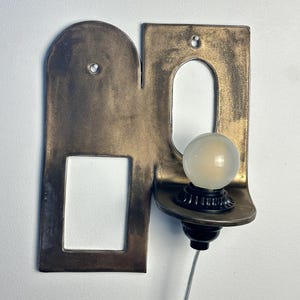 K&ouml;nnte beinhalten: Eine Wandleuchte mit einer flachen, bronzefarbenen Metallplatte, die einen bogenf&ouml;rmigen Ausschnitt und eine rechteckige &Ouml;ffnung aufweist. Eine kleine, runde, mattierte Gl&uuml;hbirne sitzt in einer schwarzen Fassung auf einer Ablage.