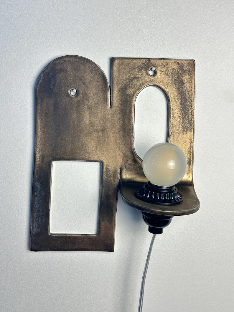 K&ouml;nnte beinhalten: Eine Wandleuchte mit einer flachen, bronzefarbenen Metallplatte, die einen bogenf&ouml;rmigen Ausschnitt und eine rechteckige &Ouml;ffnung aufweist. Eine kleine, runde, mattierte Gl&uuml;hbirne sitzt in einer schwarzen Fassung auf einer Ablage.