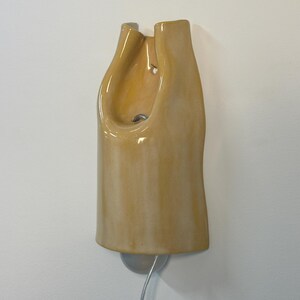 Pu&ograve; includere: Una lampada da parete con un paralume in ceramica giallo chiaro lucido caratterizzato da una forma organica, curva e allungata. L'apparecchio presenta un'apertura centrale, un foro di montaggio visibile e un cavo elettrico argentato verso il basso.