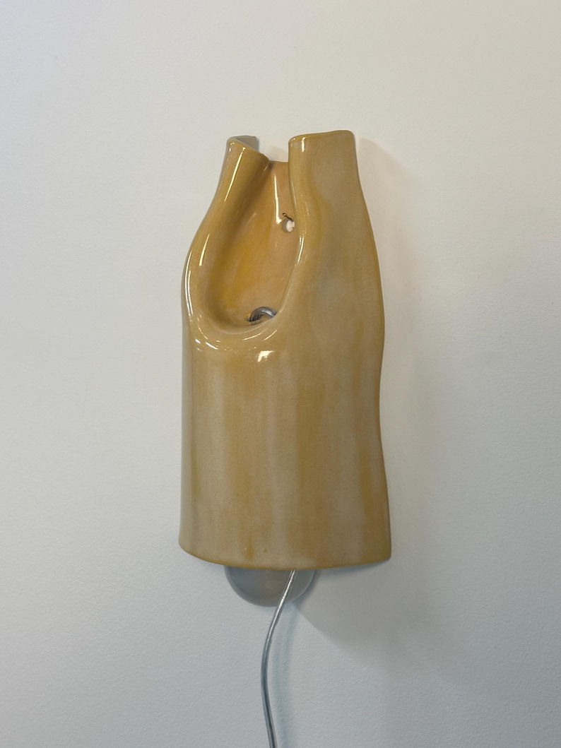 Pu&ograve; includere: Una lampada da parete con un paralume in ceramica giallo chiaro lucido caratterizzato da una forma organica, curva e allungata. L'apparecchio presenta un'apertura centrale, un foro di montaggio visibile e un cavo elettrico argentato verso il basso.