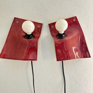 Puede incluir: Un par de l&aacute;mparas de pared con placas traseras de cer&aacute;mica roja brillante y bordes irregulares. Cada l&aacute;mpara tiene un casquillo negro central con una bombilla redonda de cristal esmerilado blanco. Un cable negro cuelga de cada l&aacute;mpara.