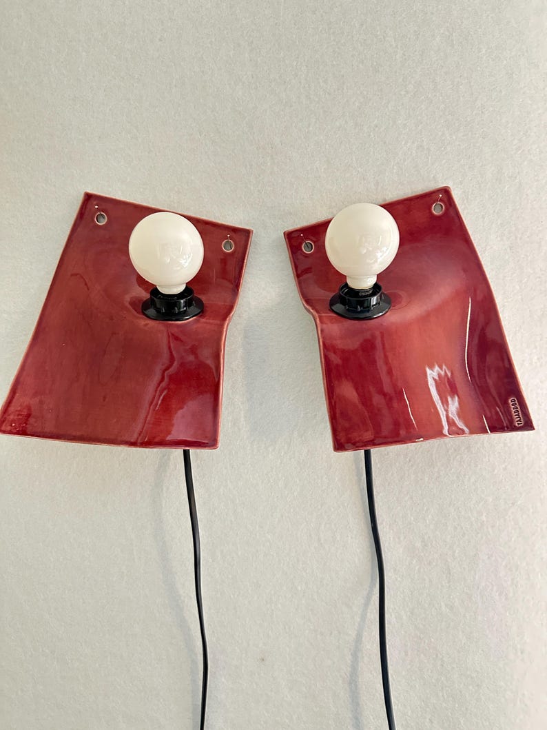 Puede incluir: Un par de l&aacute;mparas de pared con placas traseras de cer&aacute;mica roja brillante y bordes irregulares. Cada l&aacute;mpara tiene un casquillo negro central con una bombilla redonda de cristal esmerilado blanco. Un cable negro cuelga de cada l&aacute;mpara.