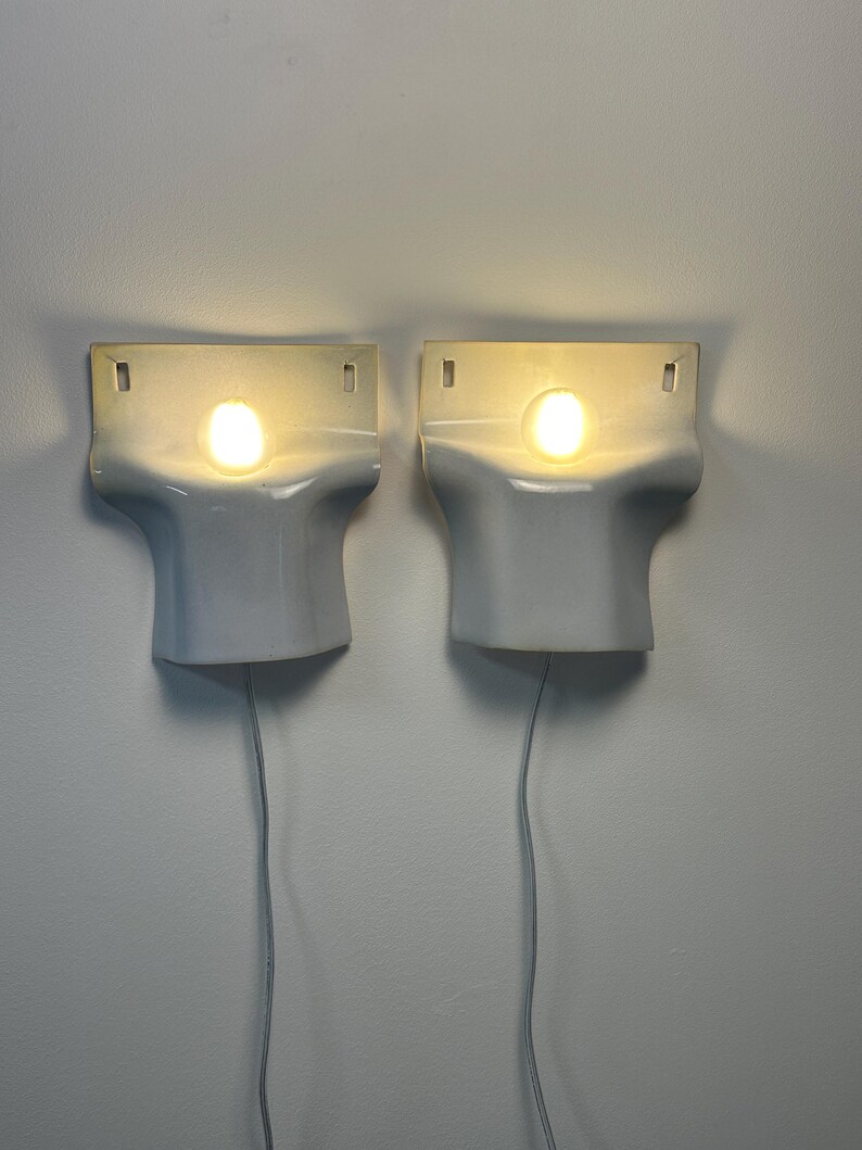 May include: A pair of white wall-mounted sconces with a smooth, curved, T-shaped design. Each fixture features a central circular light source emitting a warm glow, with two small rectangular mounting holes at the top and a thin power cord extending downward.