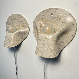May include: Two wall-mounted ceramic sconces with a speckled cream and brown finish. Each lamp features a rounded, organic shape with a central circular opening holding a frosted white glass bulb. A thin grey power cord extends from the bottom.