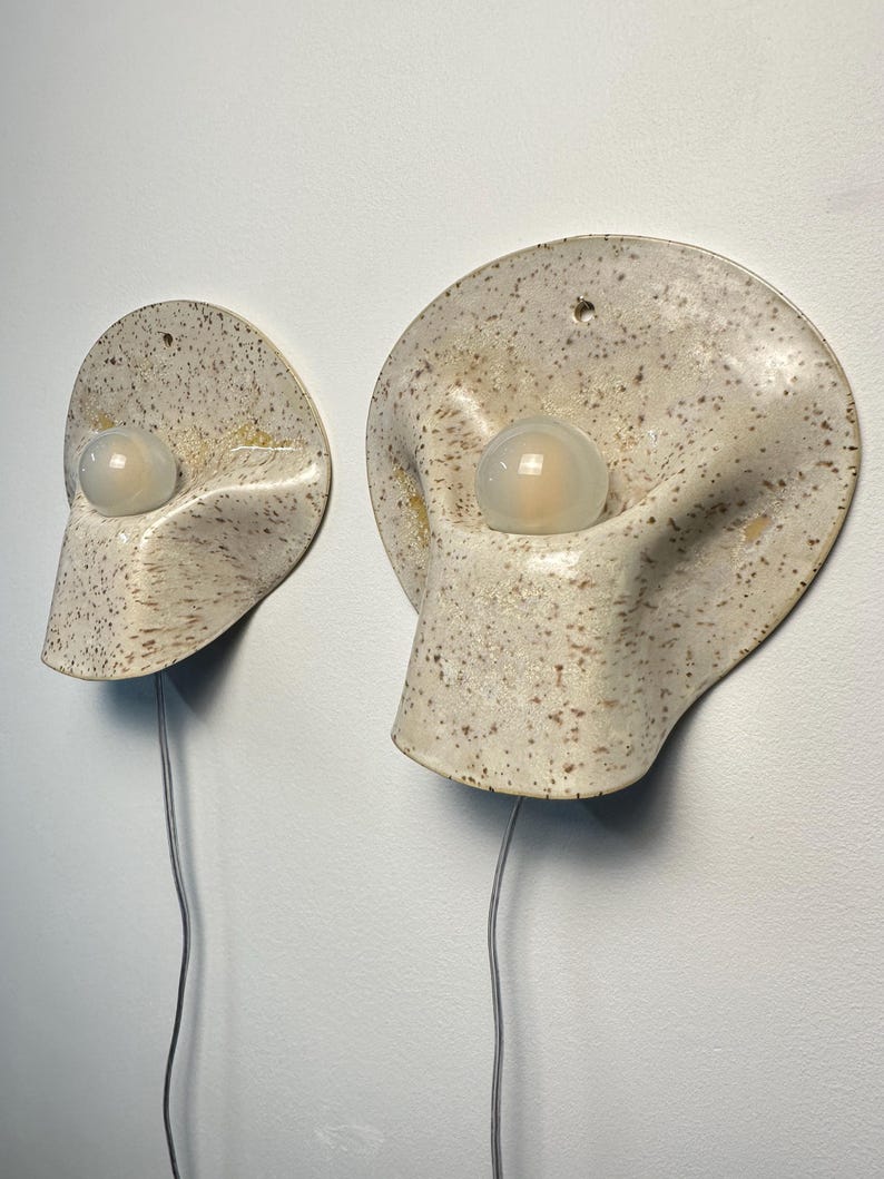 May include: Two wall-mounted ceramic sconces with a speckled cream and brown finish. Each lamp features a rounded, organic shape with a central circular opening holding a frosted white glass bulb. A thin grey power cord extends from the bottom.