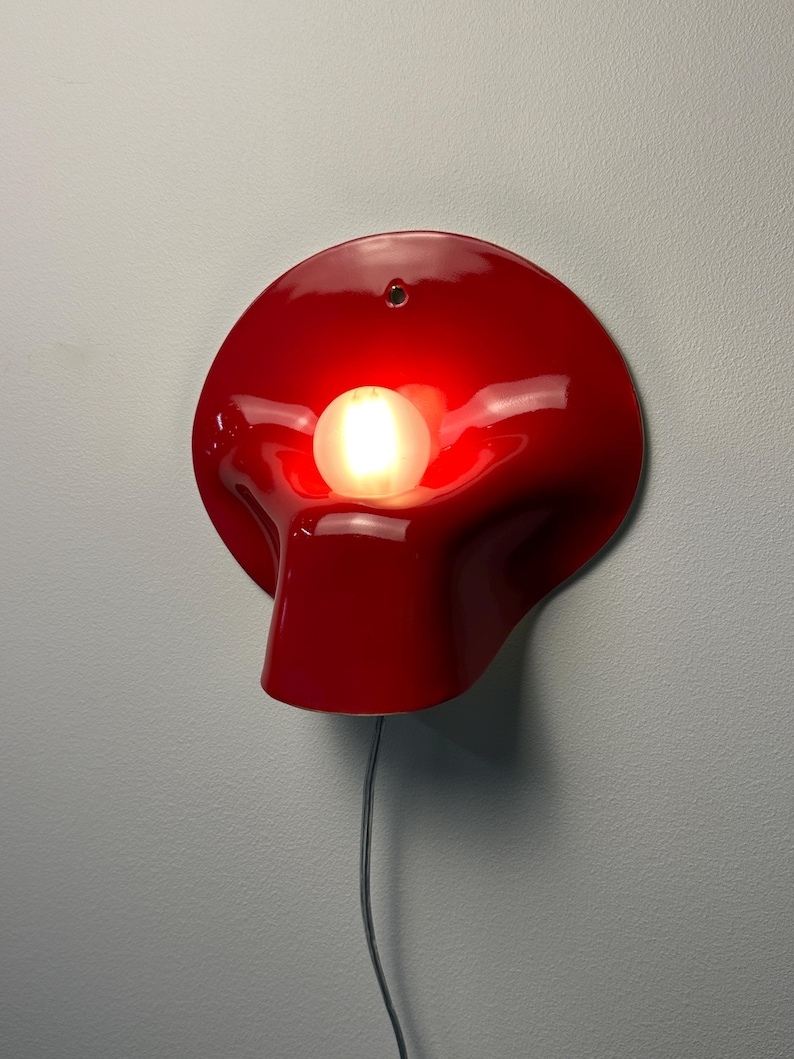 May include: A glossy red wall lamp with a curved, organic shape and a central circular opening. A single glowing white spherical light bulb is positioned within the center of the red fixture. A thin black power cord hangs from the bottom of the lamp.