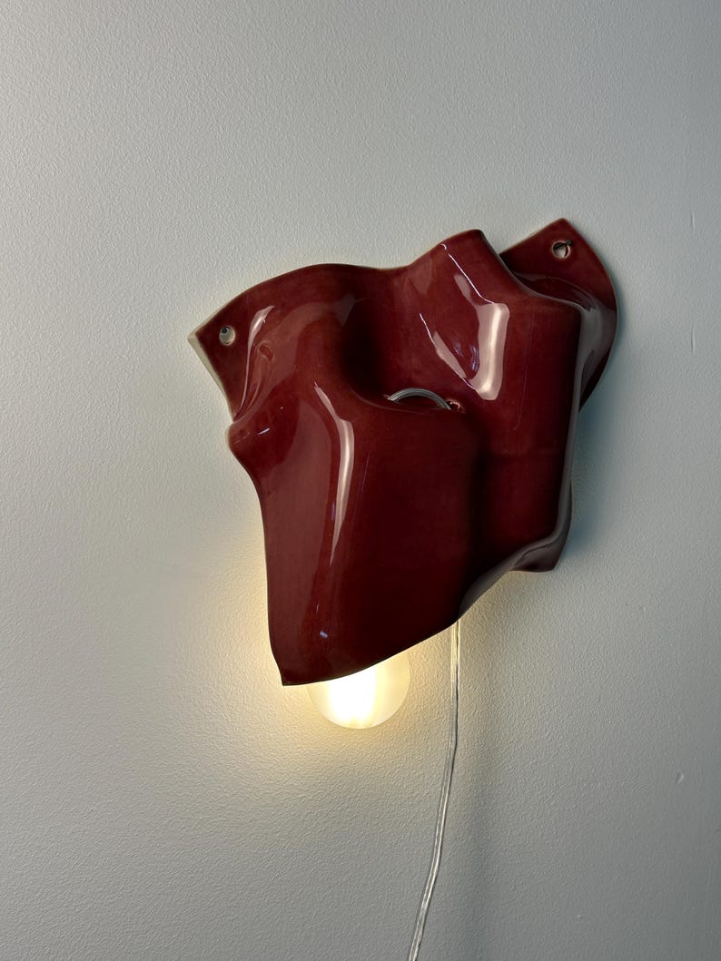 Puede incluir: Una l&aacute;mpara de pared con una pantalla de cer&aacute;mica de color rojo intenso brillante con una forma org&aacute;nica, irregular y ondulada. El aplique incluye una bombilla expuesta que emite una luz c&aacute;lida y un cable de alimentaci&oacute;n transparente.