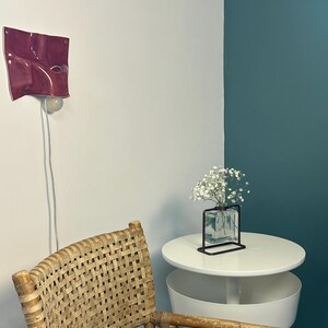 May include: A wall-mounted magenta ceramic light fixture with a wavy, folded surface and a single exposed bulb. The lamp features a white power cord hanging down. It is positioned above a woven rattan chair and a white round side table.