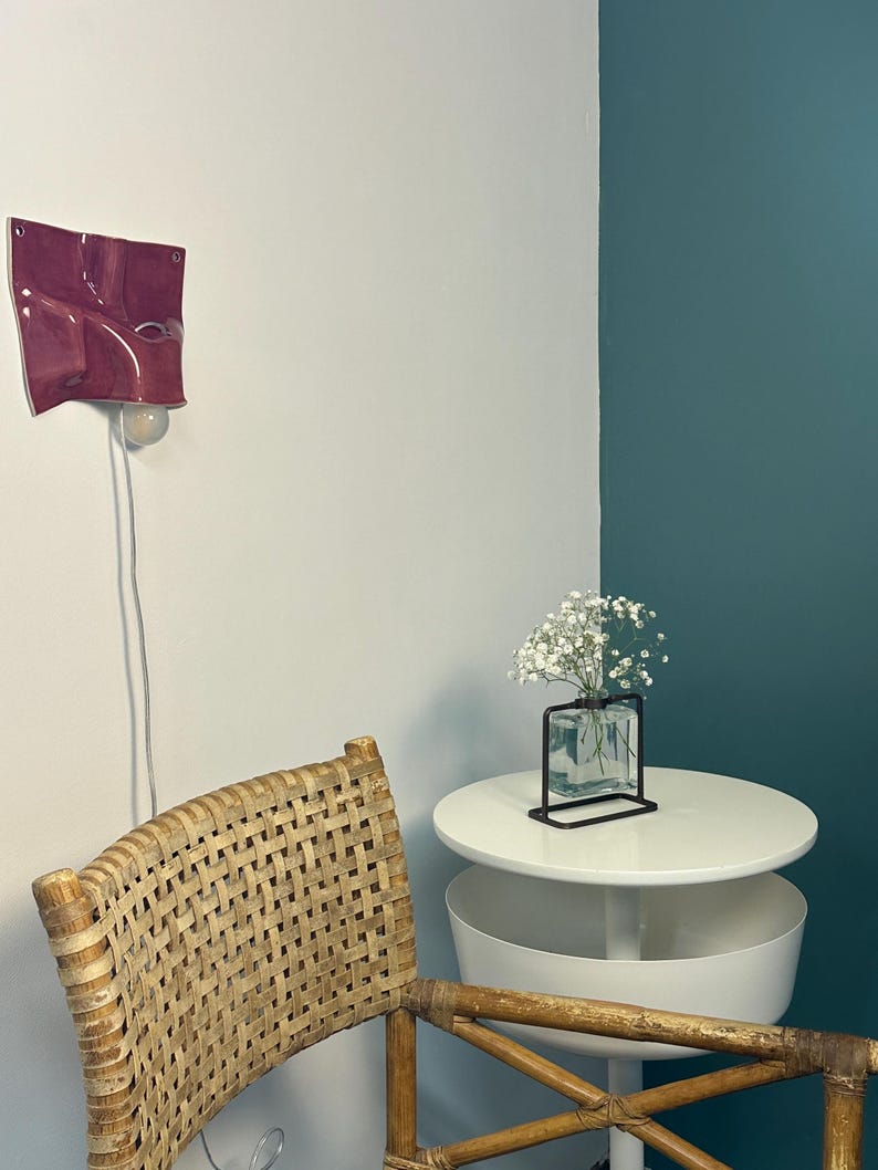 May include: A wall-mounted magenta ceramic light fixture with a wavy, folded surface and a single exposed bulb. The lamp features a white power cord hanging down. It is positioned above a woven rattan chair and a white round side table.