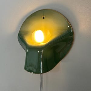 May include: A wall-mounted ceramic lamp with a glossy finish, featuring a gradient of dark green to light yellow. The curved, shell-like form houses a single glowing light bulb in the center and includes a small hole at the top for mounting.