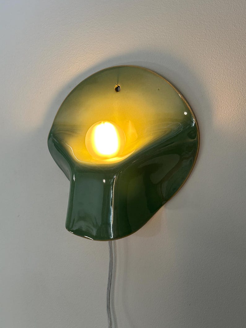 May include: A wall-mounted ceramic lamp with a glossy finish, featuring a gradient of dark green to light yellow. The curved, shell-like form houses a single glowing light bulb in the center and includes a small hole at the top for mounting.