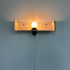 May include: A wall-mounted light fixture with a curved, speckled shade and a single exposed light bulb. The fixture has a metallic frame and is mounted on a light blue wall. The bulb is lit, casting a warm glow.