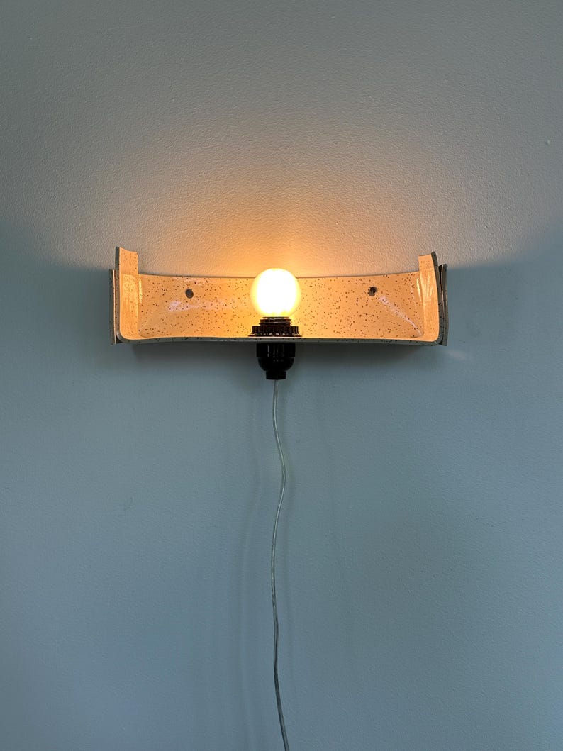 May include: A wall-mounted light fixture with a curved, speckled shade and a single exposed light bulb. The fixture has a metallic frame and is mounted on a light blue wall. The bulb is lit, casting a warm glow.