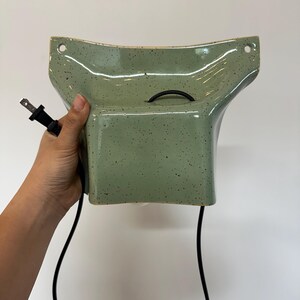 May include: A sage green ceramic wall sconce with a speckled finish and a flared, wide-top design. The fixture features two circular mounting holes at the top corners and includes a black electrical cord with a standard two-prong plug attached.