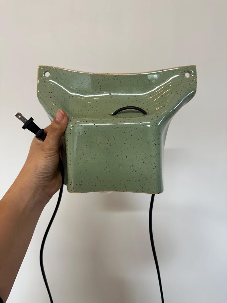 May include: A sage green ceramic wall sconce with a speckled finish and a flared, wide-top design. The fixture features two circular mounting holes at the top corners and includes a black electrical cord with a standard two-prong plug attached.