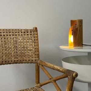 Puede incluir: Una silla con respaldo y asiento de ca&ntilde;a trenzada color canela, con estructura de bamb&uacute; marr&oacute;n claro y reposabrazos en cruz. Junto a la silla hay una peque&ntilde;a mesa blanca con una l&aacute;mpara cil&iacute;ndrica de cer&aacute;mica que emite una luz c&aacute;lida.