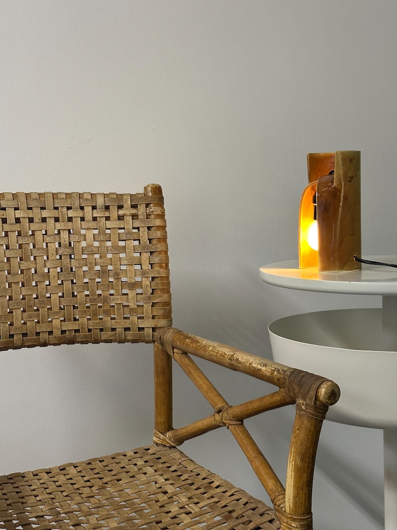 Puede incluir: Una silla con respaldo y asiento de ca&ntilde;a trenzada color canela, con estructura de bamb&uacute; marr&oacute;n claro y reposabrazos en cruz. Junto a la silla hay una peque&ntilde;a mesa blanca con una l&aacute;mpara cil&iacute;ndrica de cer&aacute;mica que emite una luz c&aacute;lida.