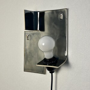 Pode incluir: Uma arandela de parede moderna com base em cer&acirc;mica em tons de cinza e preto. A arandela apresenta uma l&acirc;mpada branca e um cabo preto. O design &eacute; minimalista e art&iacute;stico, adequado para ilumina&ccedil;&atilde;o interior.