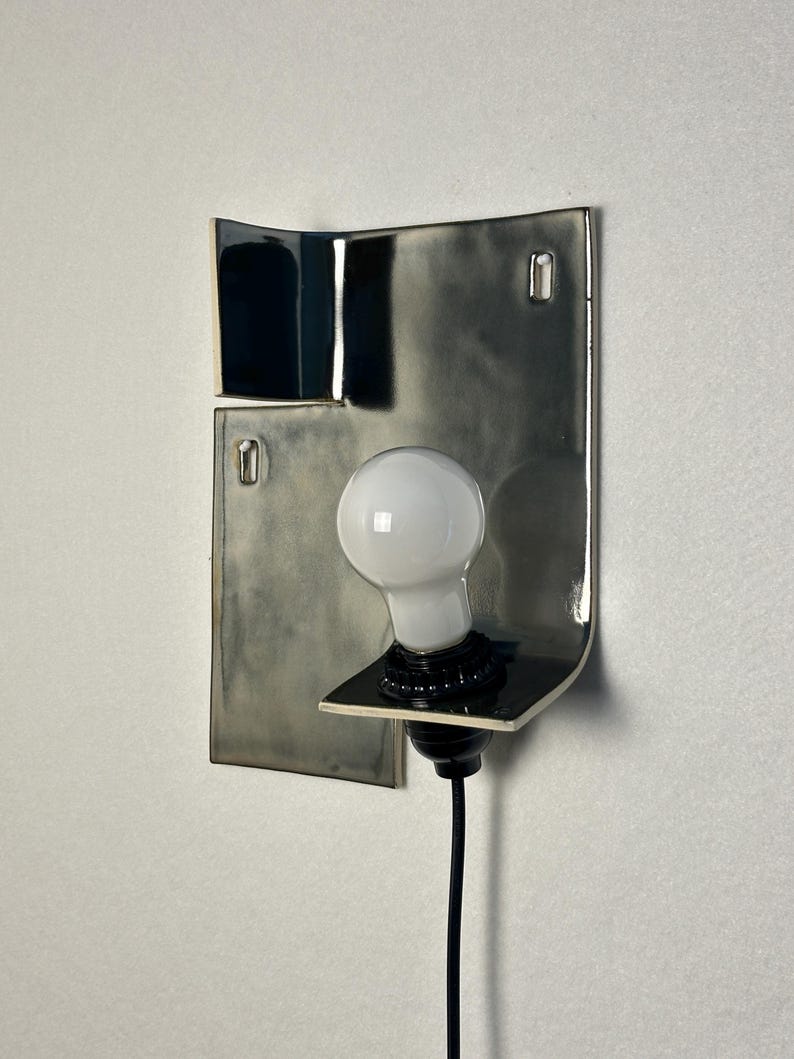 Pode incluir: Uma arandela de parede moderna com base em cer&acirc;mica em tons de cinza e preto. A arandela apresenta uma l&acirc;mpada branca e um cabo preto. O design &eacute; minimalista e art&iacute;stico, adequado para ilumina&ccedil;&atilde;o interior.