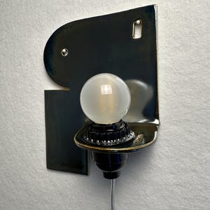 May include: A wall-mounted sconce featuring a glossy, dark navy blue ceramic backplate with an irregular, organic shape. A black socket holds a single, round, frosted glass light bulb. A thin, clear electrical cord extends downwards from the base.