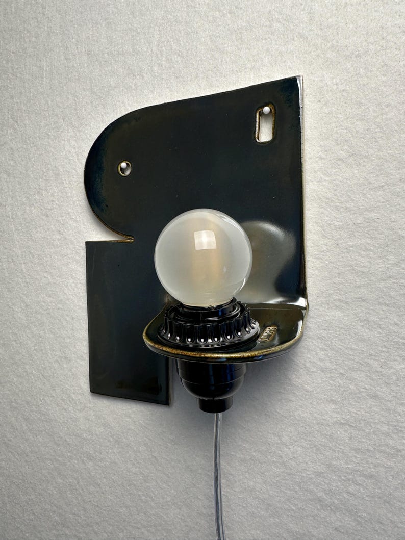 May include: A wall-mounted sconce featuring a glossy, dark navy blue ceramic backplate with an irregular, organic shape. A black socket holds a single, round, frosted glass light bulb. A thin, clear electrical cord extends downwards from the base.