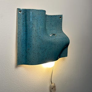 May include: A wall-mounted ceramic light fixture with a glossy turquoise glaze and dark speckles. The sculptural form features an asymmetrical, curved shape with a central opening for a light bulb, a small metal ring, and a clear power cord with a switch.