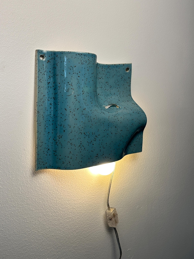 May include: A wall-mounted ceramic light fixture with a glossy turquoise glaze and dark speckles. The sculptural form features an asymmetrical, curved shape with a central opening for a light bulb, a small metal ring, and a clear power cord with a switch.