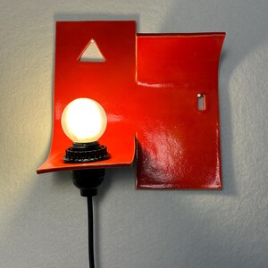 May include: A contemporary red wall light with a distinctive design. It has a bright, round bulb and a black cable. The red fixture incorporates a triangular cut-out and a keyhole shape, adding an artistic touch to the lighting.