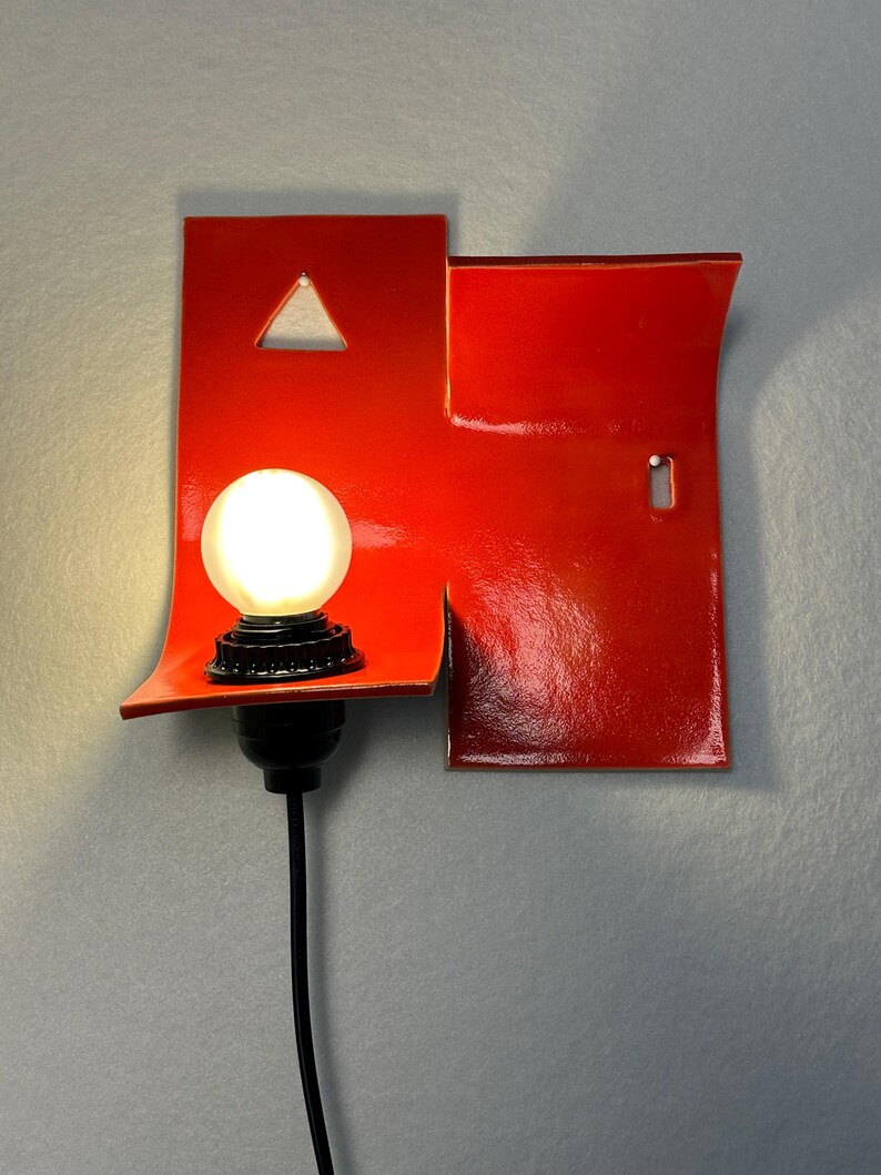May include: A contemporary red wall light with a distinctive design. It has a bright, round bulb and a black cable. The red fixture incorporates a triangular cut-out and a keyhole shape, adding an artistic touch to the lighting.