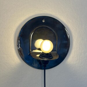 May include: A circular, dark blue ceramic wall sconce featuring a glossy finish and a central arched cutout. A small, round, glowing light bulb is mounted within the cutout, and a black power cord extends downward from the base of the fixture.