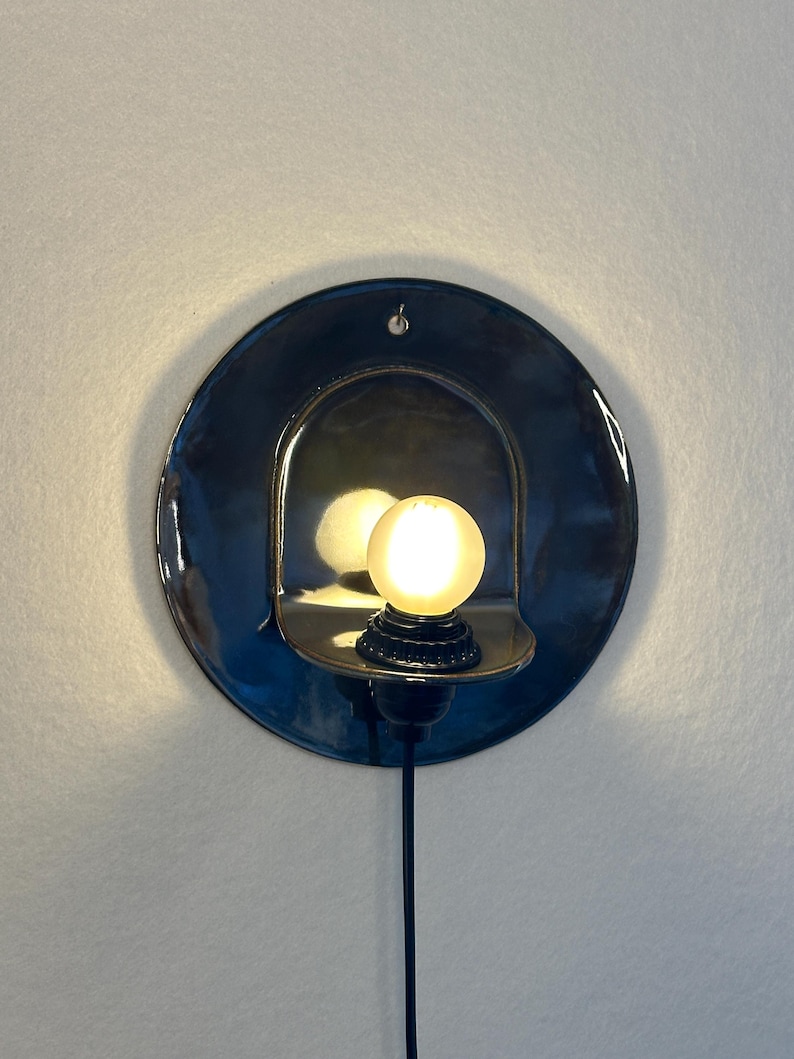May include: A circular, dark blue ceramic wall sconce featuring a glossy finish and a central arched cutout. A small, round, glowing light bulb is mounted within the cutout, and a black power cord extends downward from the base of the fixture.