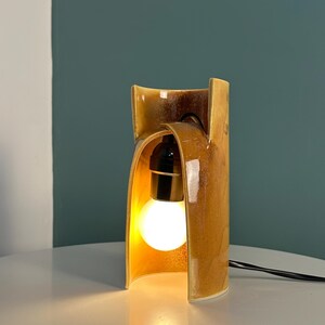Puede incluir: Una l&aacute;mpara de mesa de cer&aacute;mica con un esmalte brillante de color &aacute;mbar marr&oacute;n moteado. La estructura presenta una forma curva similar a un arco que encierra parcialmente un casquillo negro central con una bombilla blanca redonda.