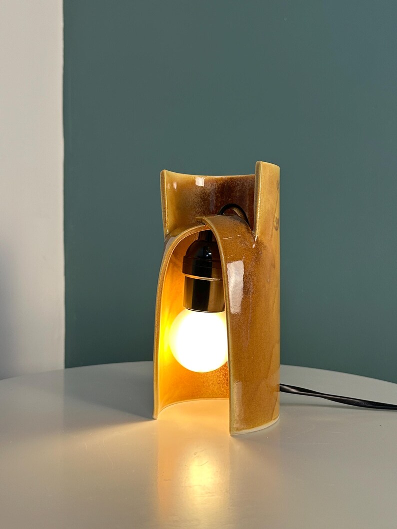 Puede incluir: Una l&aacute;mpara de mesa de cer&aacute;mica con un esmalte brillante de color &aacute;mbar marr&oacute;n moteado. La estructura presenta una forma curva similar a un arco que encierra parcialmente un casquillo negro central con una bombilla blanca redonda.
