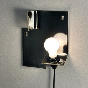 Pode incluir: Uma lumin&aacute;ria de parede moderna com um design geom&eacute;trico preto. A lumin&aacute;ria apresenta um detalhe met&aacute;lico curvo e uma &uacute;nica l&acirc;mpada iluminada. A lumin&aacute;ria &eacute; montada em uma parede cinza claro, mostrando sua est&eacute;tica minimalista.