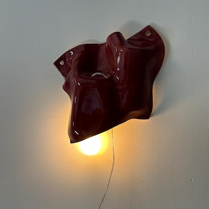 Puede incluir: Una l&aacute;mpara de pared con una carcasa de cer&aacute;mica o pl&aacute;stico de color rojo oscuro brillante, con formas org&aacute;nicas y contornos irregulares. Una bombilla de luz c&aacute;lida est&aacute; situada en la parte inferior, emitiendo un brillo suave y un cable.