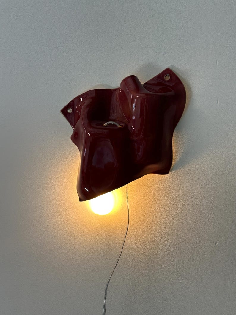Puede incluir: Una l&aacute;mpara de pared con una carcasa de cer&aacute;mica o pl&aacute;stico de color rojo oscuro brillante, con formas org&aacute;nicas y contornos irregulares. Una bombilla de luz c&aacute;lida est&aacute; situada en la parte inferior, emitiendo un brillo suave y un cable.