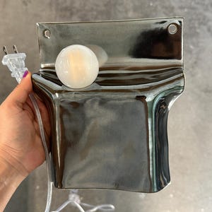 Peut inclure: Une lampe murale dot&eacute;e d'une plaque arri&egrave;re rectangulaire incurv&eacute;e en m&eacute;tal argent&eacute; r&eacute;fl&eacute;chissant, avec deux trous de fixation circulaires en haut. Une ampoule ronde en verre d&eacute;poli blanc est fix&eacute;e sur la surface avant, reli&eacute;e &agrave; un cordon &eacute;lectrique transparent et une prise.
