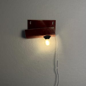 Może przedstawiać: Lampa ścienna z prostokątną, ciemnoczerwoną, zakrzywioną metalową płytą tylną. Pojedyncza, mała, okrągła, świecąca żar&oacute;wka jest przymocowana do dolnej krawędzi zakrzywionego metalowego elementu. Cienki, przezroczysty przew&oacute;d elektryczny zwisa z oprawy.