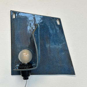 May include: A modern wall sconce with a blue glazed ceramic shade. The shade has a unique, angular shape and features a curved metal accent. A frosted bulb sits in a black socket, and a silver cord extends from the base.
