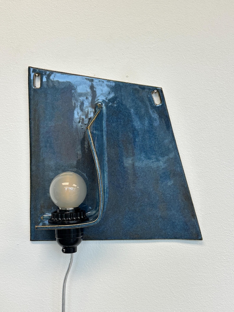 May include: A modern wall sconce with a blue glazed ceramic shade. The shade has a unique, angular shape and features a curved metal accent. A frosted bulb sits in a black socket, and a silver cord extends from the base.