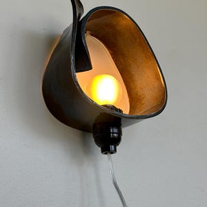 May include: A unique wall sconce with a dark, curved metal shade. The interior of the shade is a warm gold color, and it houses a lit light bulb. The sconce is mounted on a light-colored wall, with a black base and a silver cord.