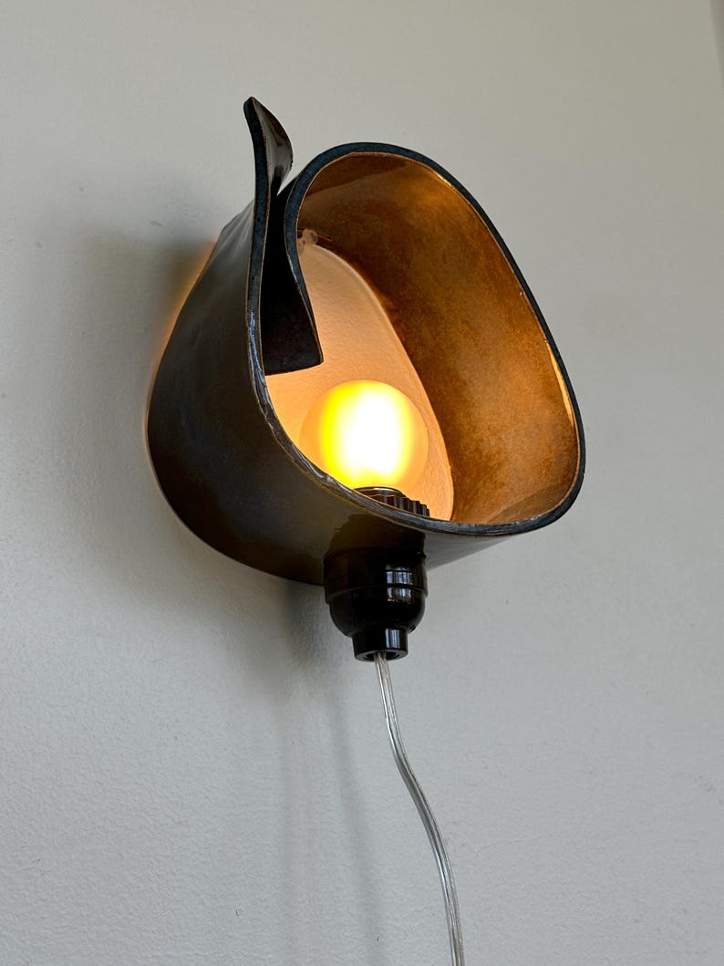 May include: A unique wall sconce with a dark, curved metal shade. The interior of the shade is a warm gold color, and it houses a lit light bulb. The sconce is mounted on a light-colored wall, with a black base and a silver cord.